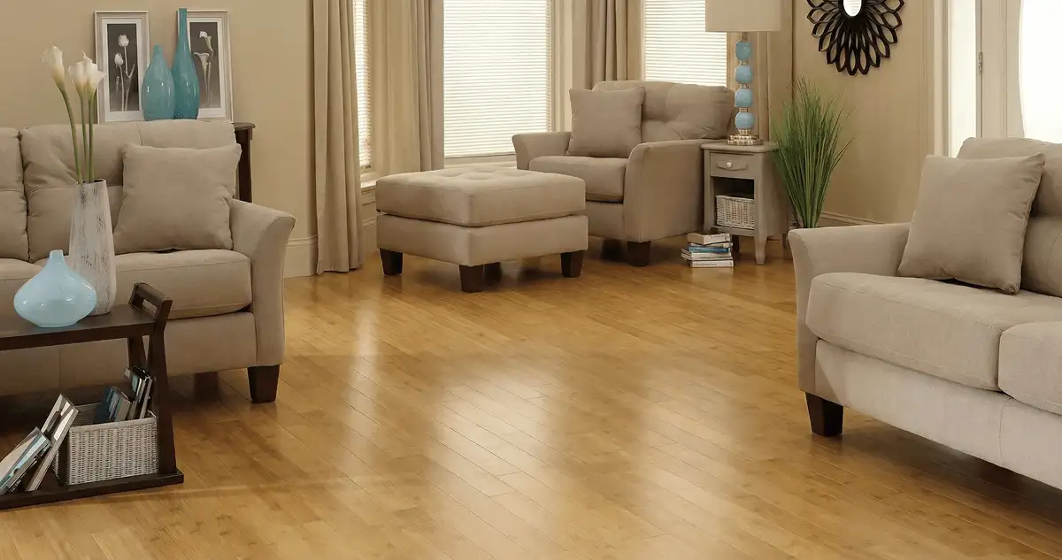 A living room with professionally installed bamboo flooring, beige sofas, a matching ottoman, a side table with a blue lamp, vases, framed pictures, and large windows with blinds and curtains in Clearwater, FL